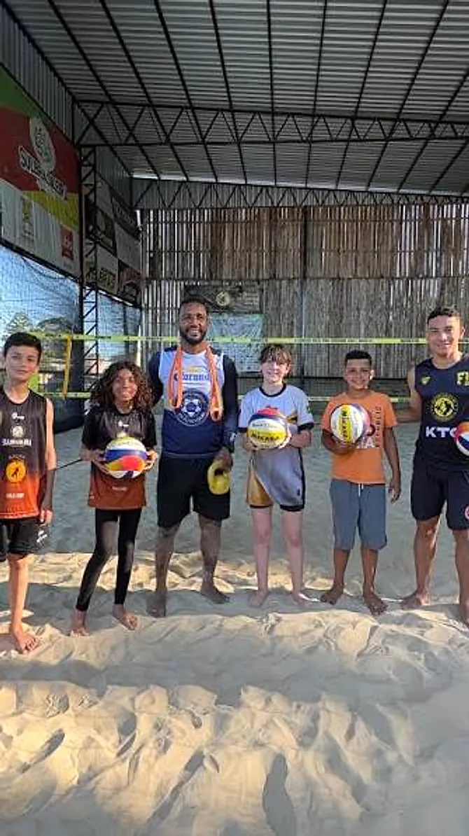 Professor com a turma de crianças na areia, cada uma com sua bola de vôlei
